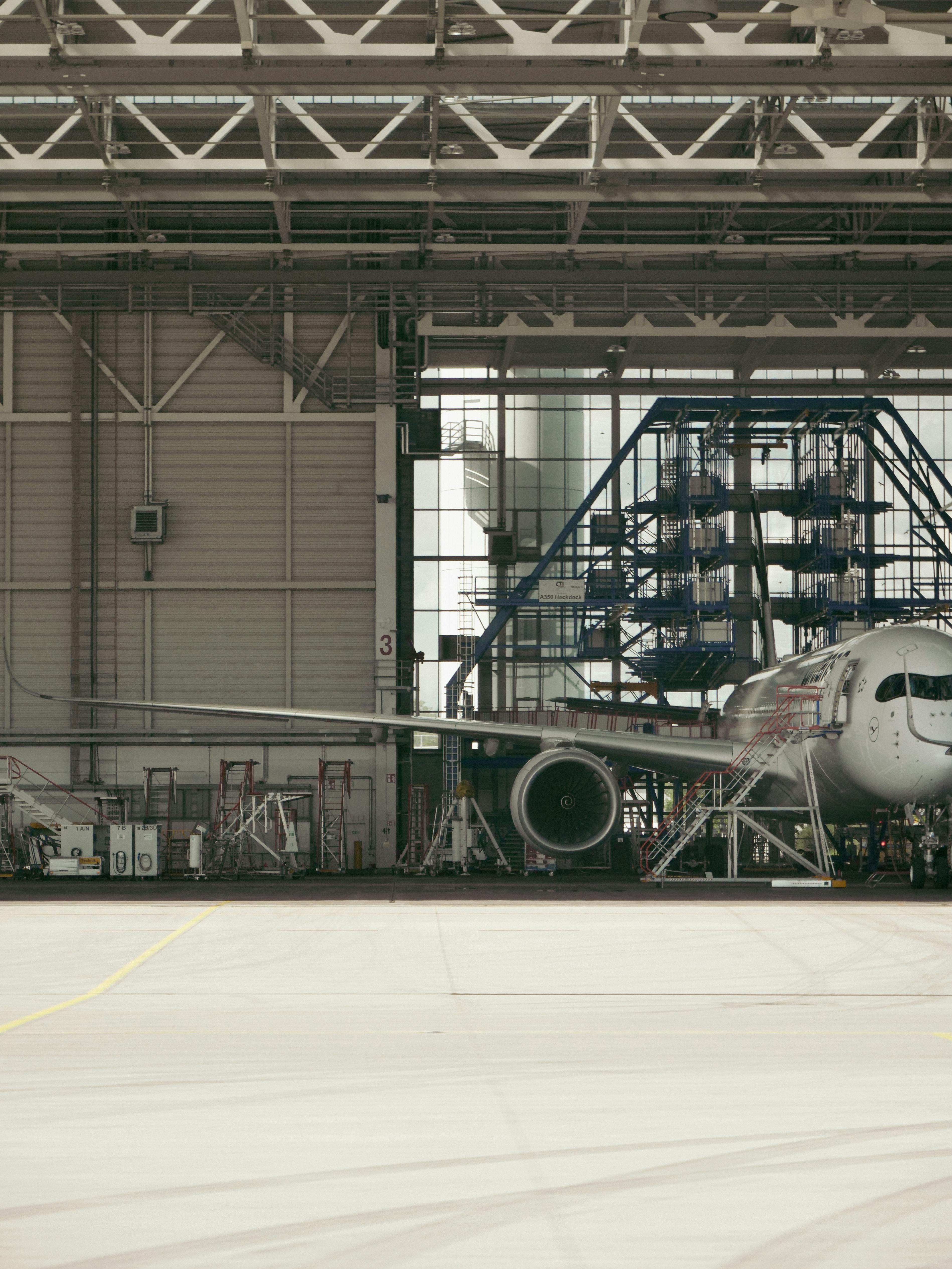 Teilansicht eines großen Verkehrsflugzeugs in einer Hangarhalle. Rechts ist die Flugzeugnase zu sehen, die Tragfläche verläuft quer durchs Bild mit einem Triebwerk darunter. Im Hintergrund stehen Wartungsgerüste, Leitern und industrielle Hallenstrukturen.