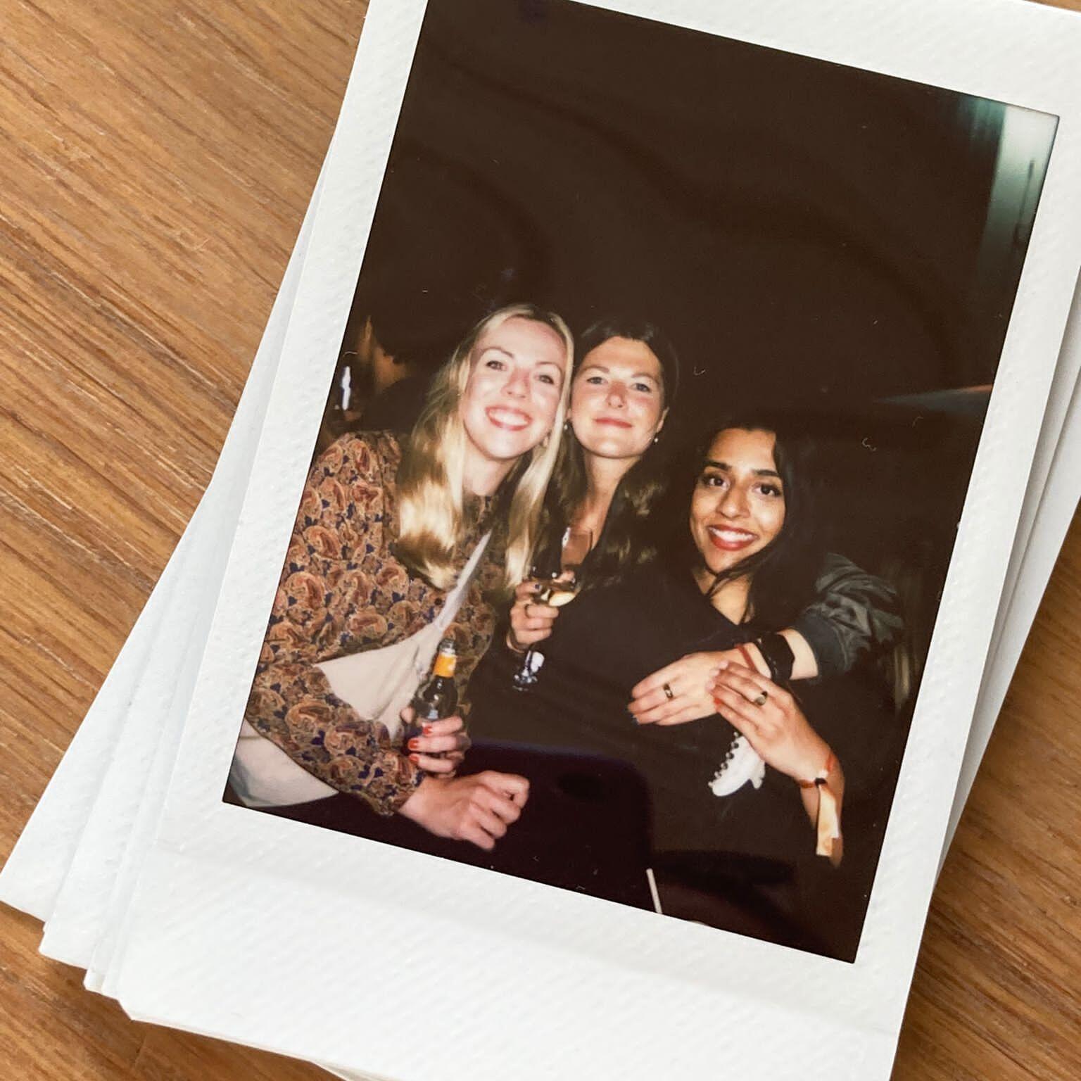 Ein Stapel Sofortbild-Fotos liegt auf einer Holzoberfläche. Das oberste Foto zeigt mehrere Personen, die gemeinsam feiern und Getränke in der Hand halten.