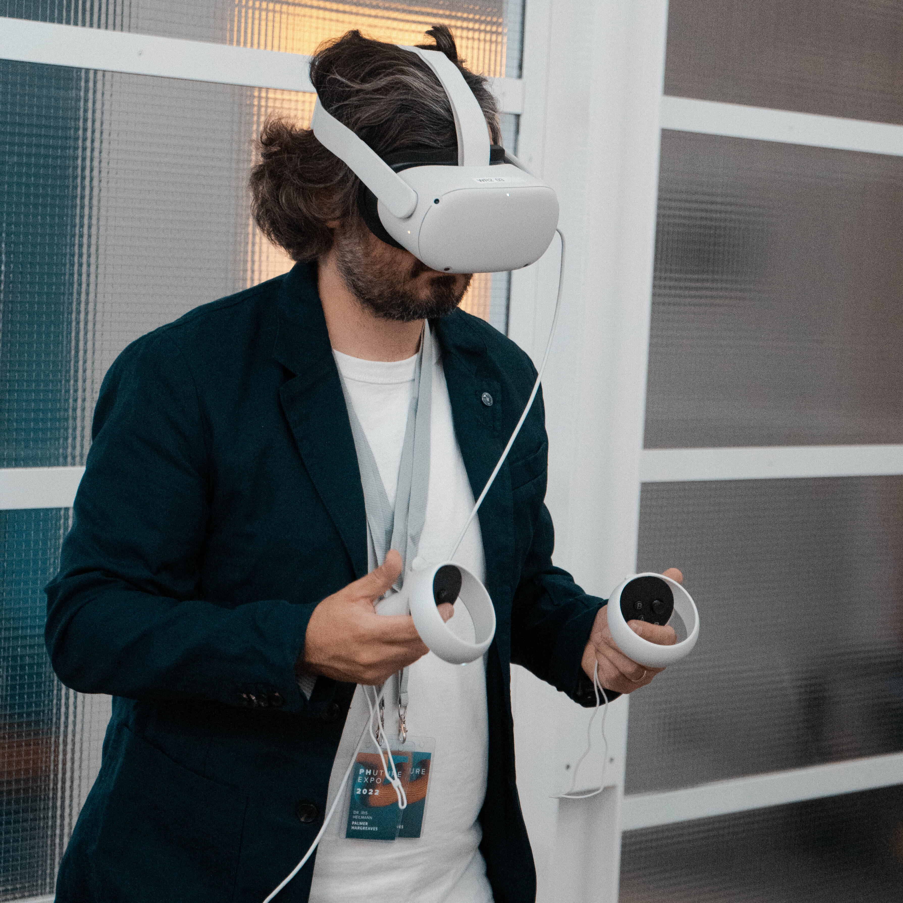 Person steht in einem Raum und trägt ein Virtual-Reality-Headset sowie zwei Controller in den Händen. Die Person ist leger gekleidet und trägt ein Namensschild um den Hals. Im Hintergrund sind Glaswände und ein Stuhl zu sehen.