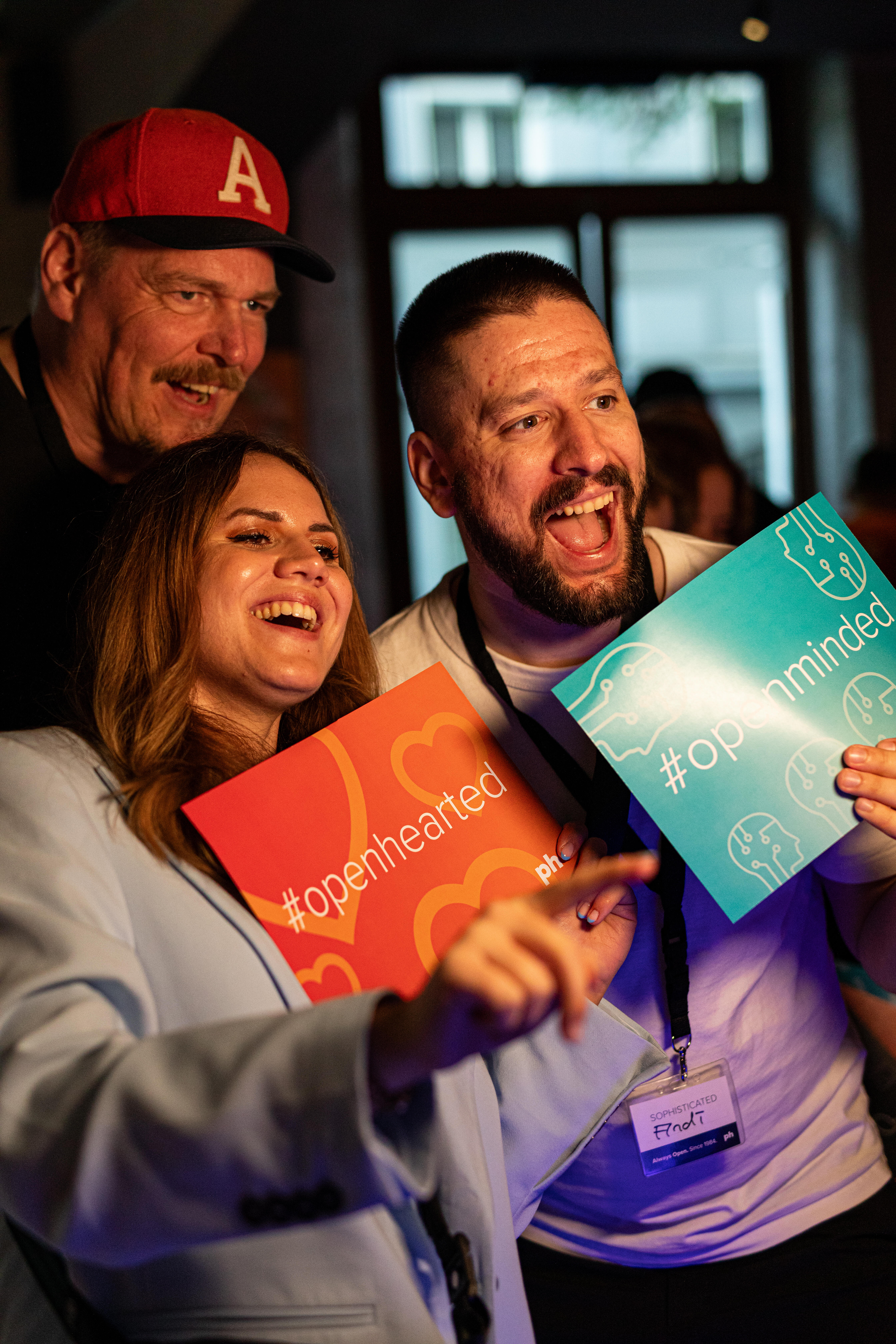 Drei Personen stehen zusammen und halten bunte Schilder mit den Aufschriften „#openhearted“ und „#openminded“ in die Kamera. Im Hintergrund ist ein Fenster zu sehen, die Stimmung wirkt fröhlich und locker.