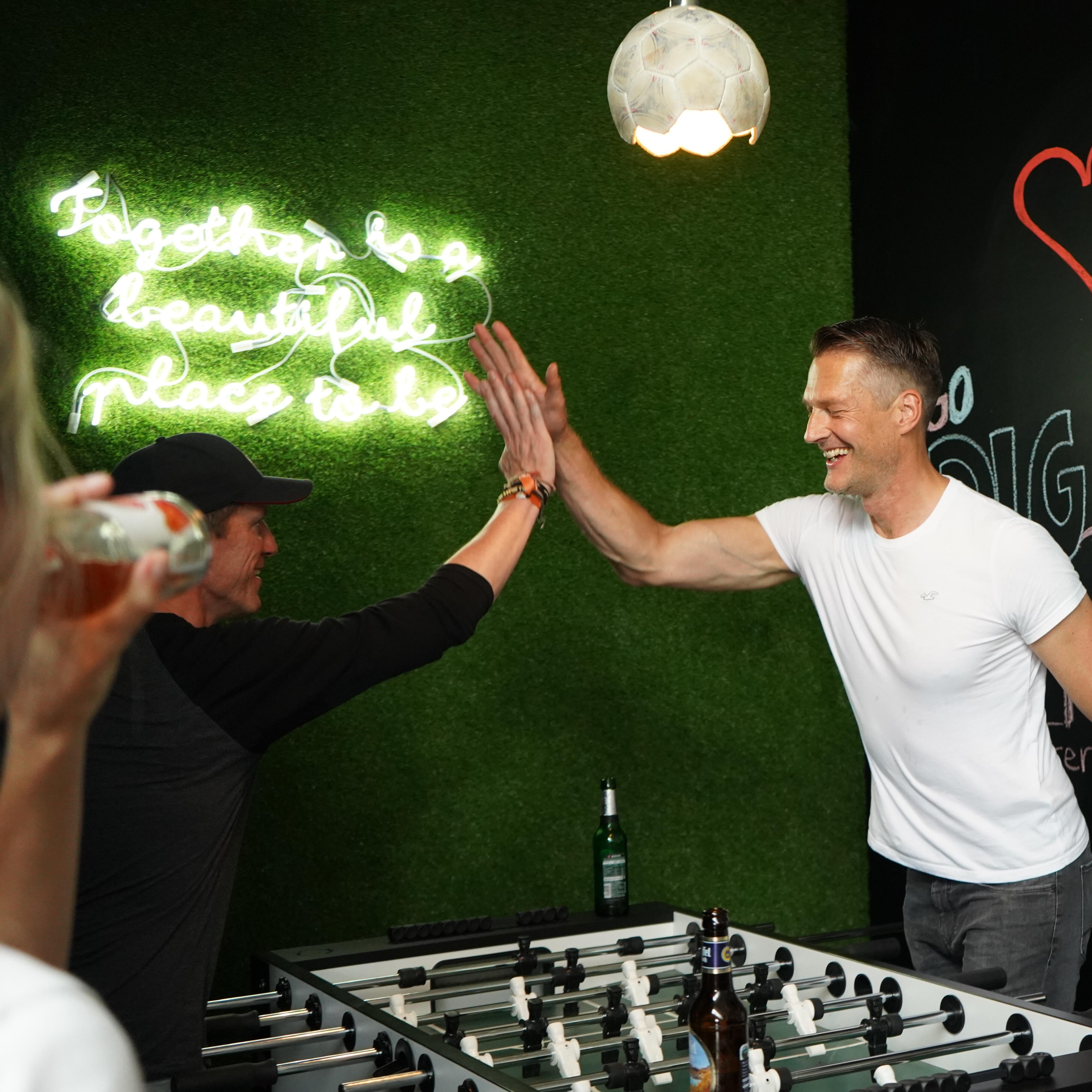 Zwei Personen geben sich ein High-Five an einem Kickertisch in einem Raum mit grüner Wand und Neon-Schriftzug. Im Hintergrund eine Wand mit Kreidezeichnungen und einem roten Herz. Weitere Personen stehen am Rand des Bildes.