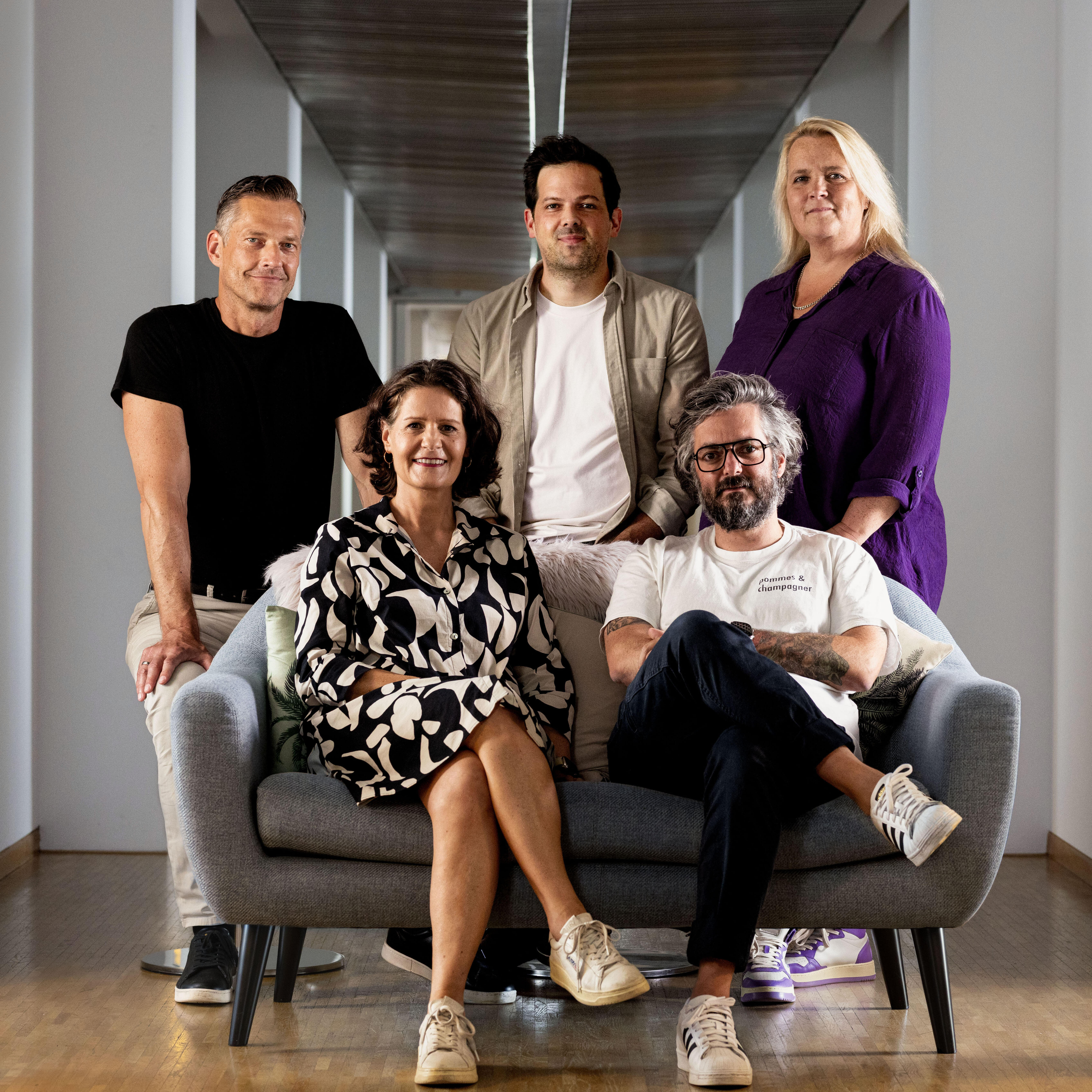 Fünf Personen sitzen und stehen auf und um ein graues Sofa in einem modernen, hellen Flur mit Holzfußboden. Die Gruppe ist leger gekleidet und blickt in Richtung Kamera.