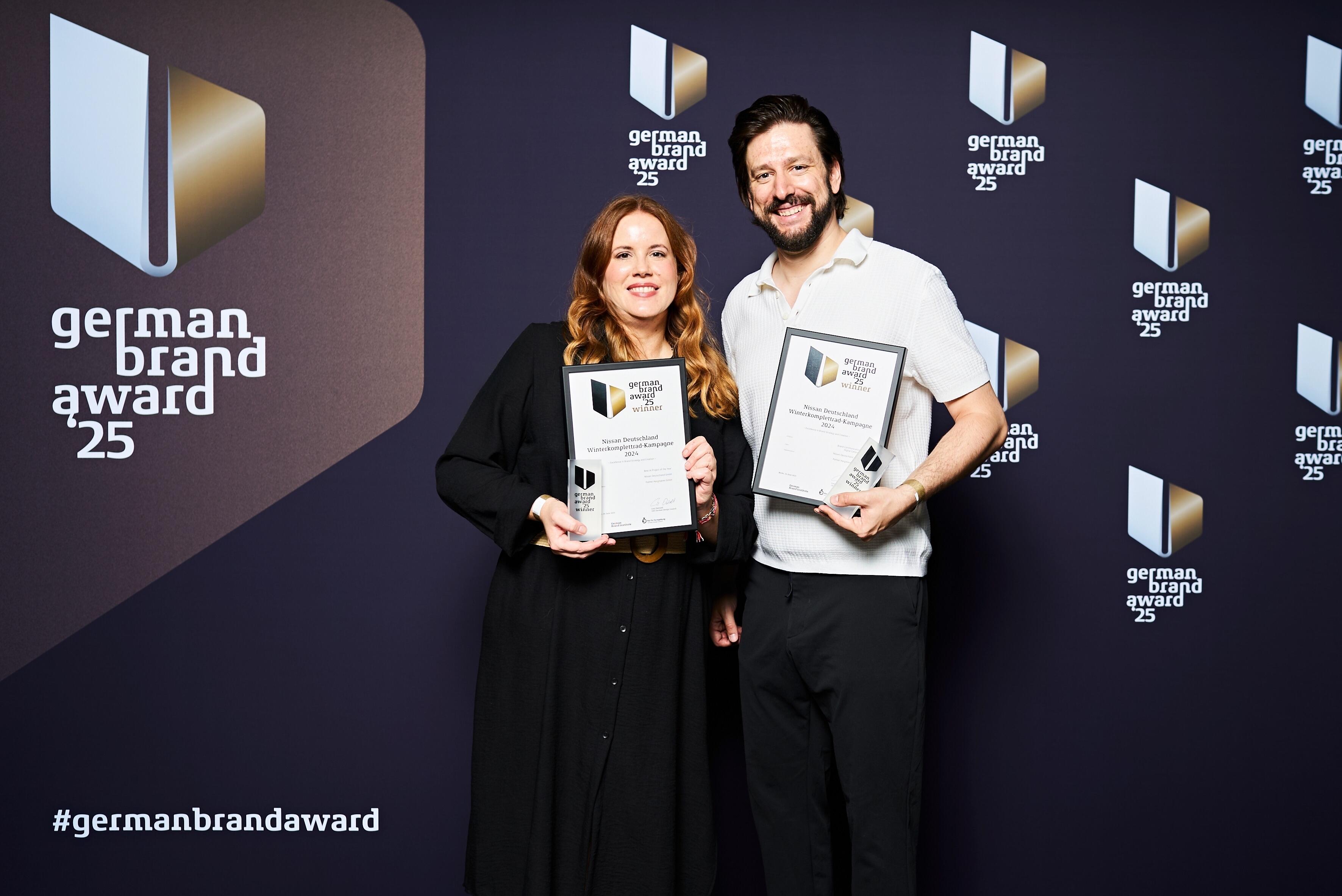 Zwei Personen stehen vor einer Fotowand mit dem Logo und Schriftzug „german brand award 25“. Beide halten eine Auszeichnung in den Händen und lächeln in die Kamera. Die Wand ist dunkel und mit weiteren Logos versehen.