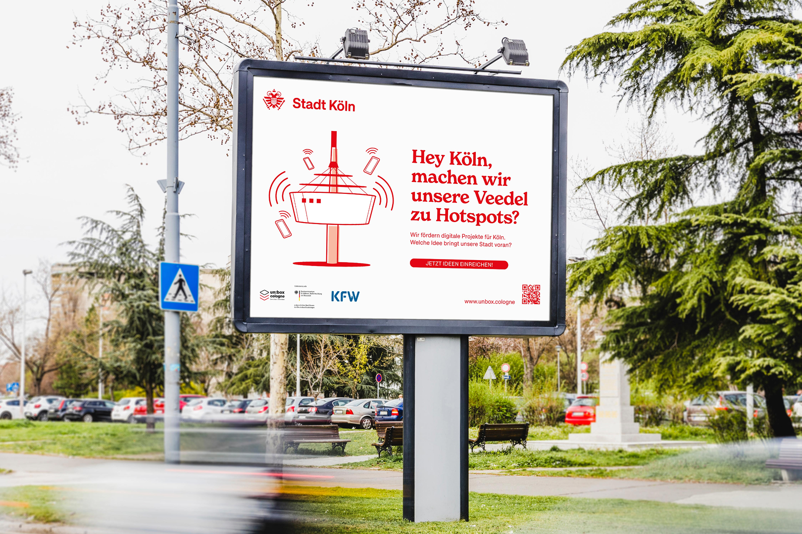Großes Werbeplakat der Stadt Köln im Freien. Auf dem Plakat steht: „Hey Köln, machen wir unsere Veedel zu Hotspots?“ und zeigt eine stilisierte Darstellung des Kölner Fernsehturms mit WLAN-Symbolen. Im Hintergrund sind Autos, Bäume und ein Park zu sehen.