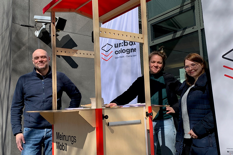Mehrere Personen stehen draußen neben einem mobilen Holzstand mit rotem Dach und der Aufschrift „Meinungs Mobil“. Im Hintergrund sind ein Banner mit dem Logo „unbox cologne“ und ein modernes Gebäude zu sehen.
