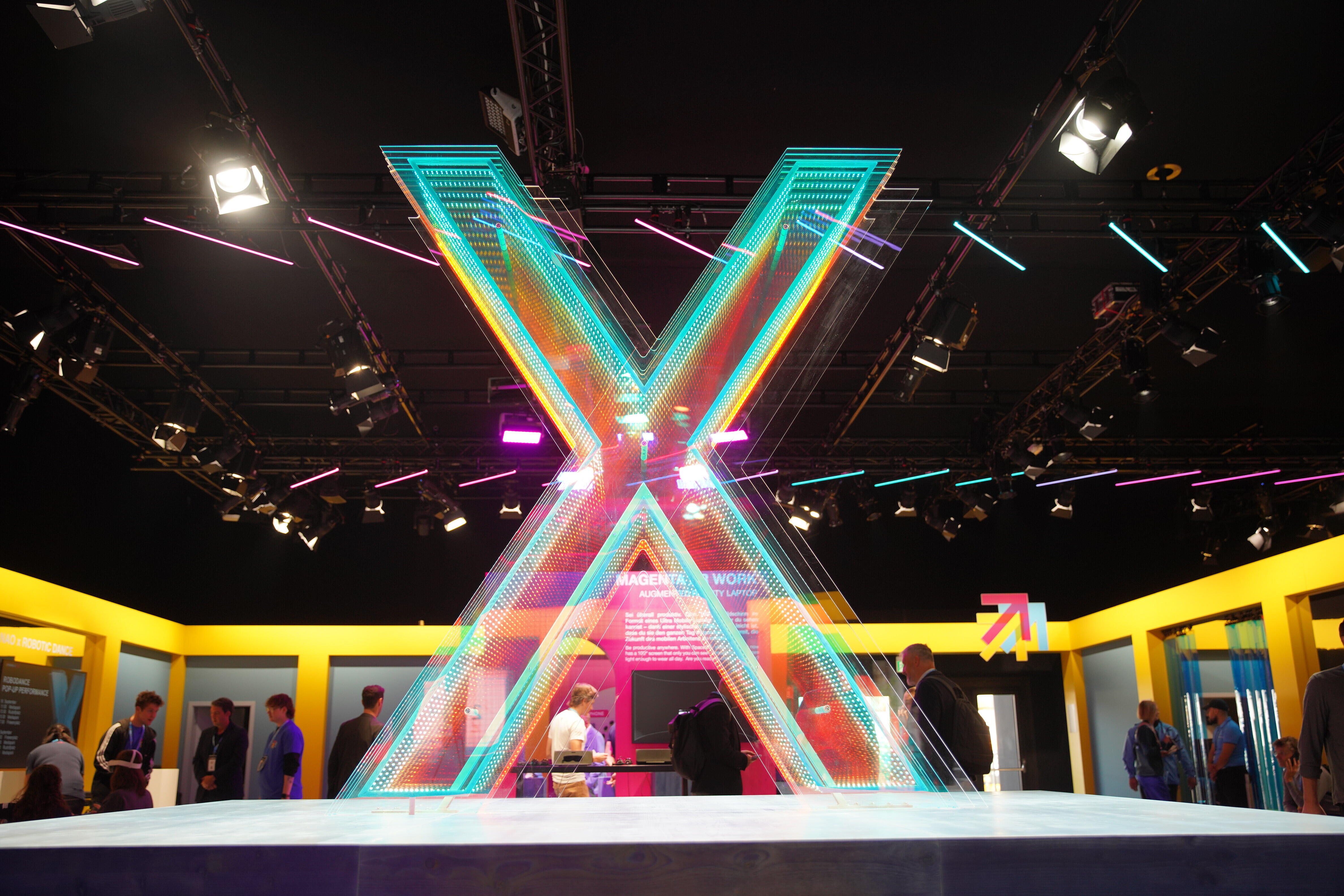 Große, leuchtende X-Skulptur aus bunten Neonlichtern steht im Mittelpunkt einer modernen Messehalle. Um die Skulptur herum sind Menschen und gelb umrahmte Stände zu sehen. Die Decke ist mit weiteren Neonröhren und Scheinwerfern ausgestattet.