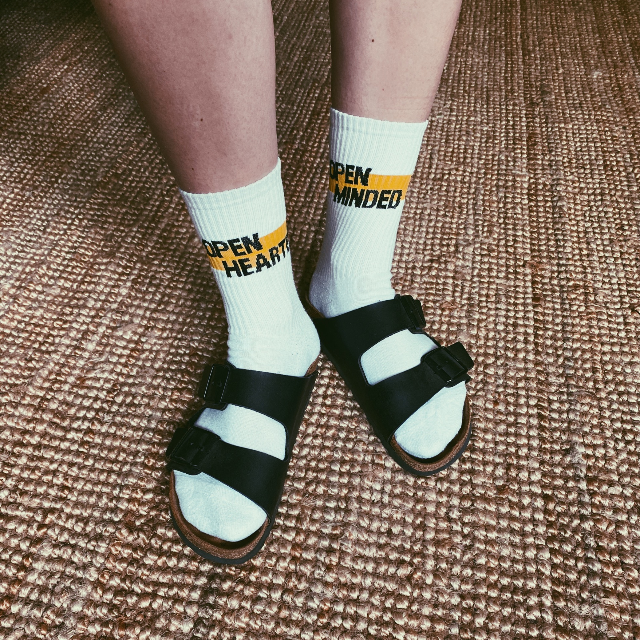 Person steht auf einem geflochtenen Teppich und trägt weiße Socken mit gelbem Streifen und den Aufschriften „OPEN HEARTED“ und „OPEN MINDED“, dazu schwarze Sandalen.