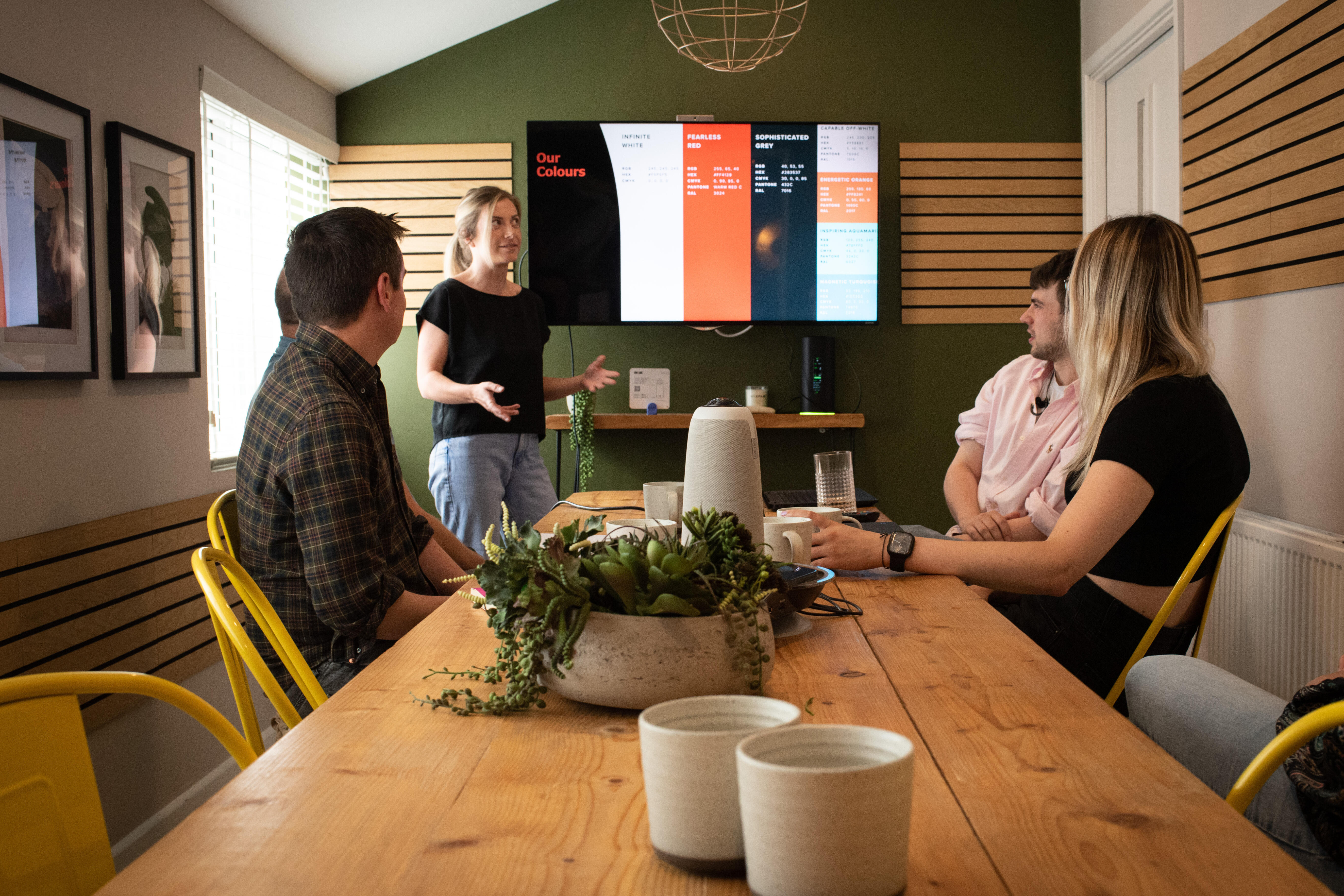Vier Personen sitzen und stehen um einen Holztisch in einem modernen Besprechungsraum. Auf dem Tisch stehen Pflanzen und Tassen. Im Hintergrund ist ein großer Bildschirm mit einer Präsentation zu sehen. Die Atmosphäre wirkt konzentriert und freundlich.