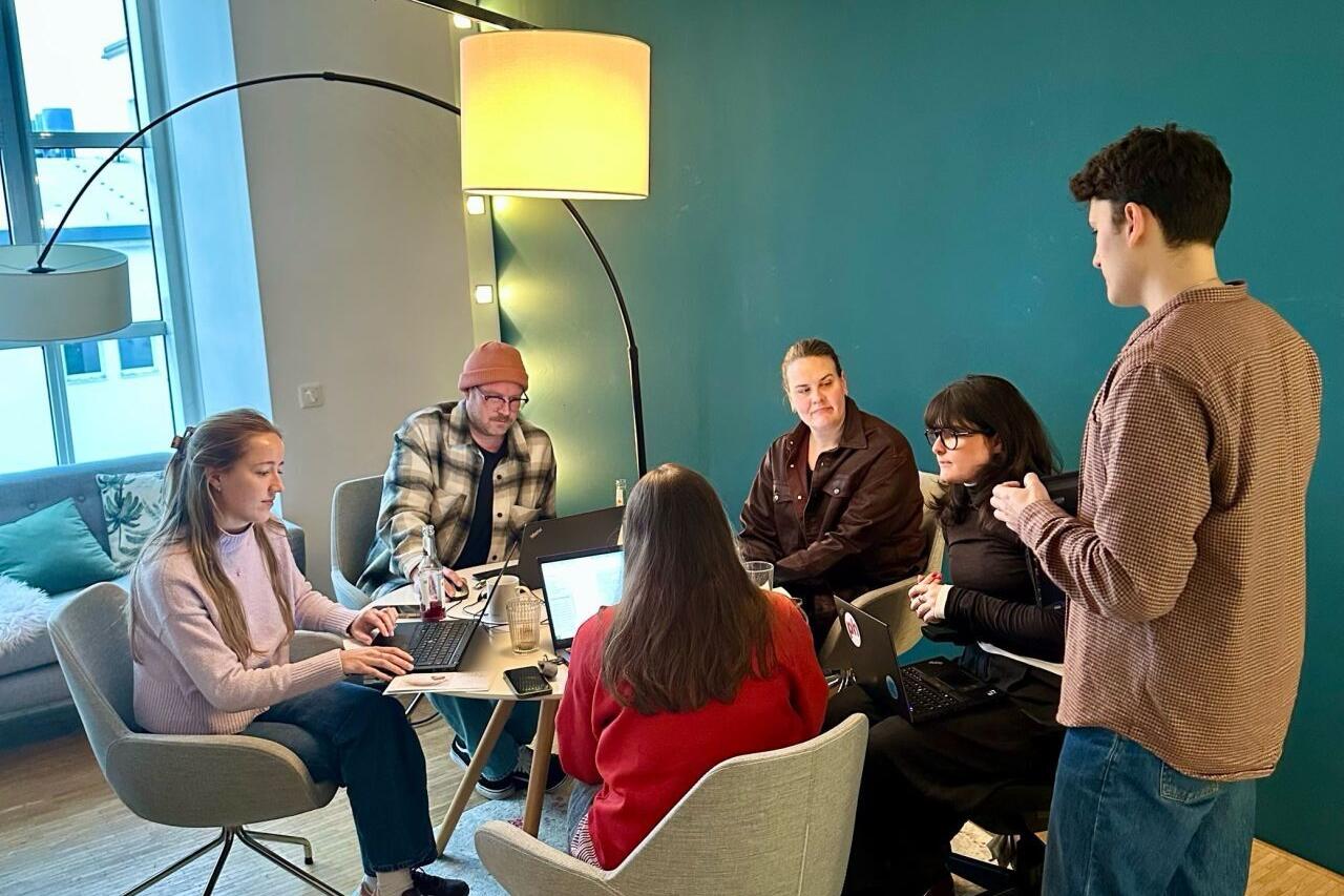 Sechs Personen sitzen und stehen um einen kleinen runden Tisch in einem modernen Büro mit türkisfarbener Wand. Mehrere Laptops stehen auf dem Tisch, und die Gruppe arbeitet gemeinsam, umgeben von Sesseln, einer Stehlampe und einem Fenster mit Tageslicht. Die Atmosphäre wirkt konzentriert und entspannt.