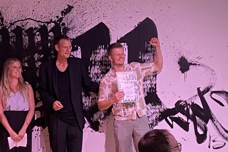 Mehrere Personen sitzen und stehen in einem Raum mit Holzfußboden. Im Vordergrund steht eine Person mit dem Rücken zur Kamera, die anderen Personen sitzen auf Stühlen und hören aufmerksam zu. Die Atmosphäre wirkt wie bei einer Präsentation oder einem Workshop.