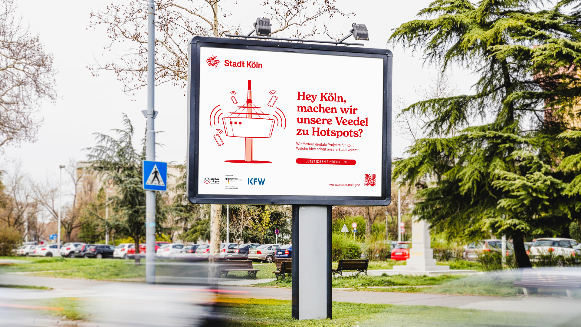 Großes Werbeplakat der Stadt Köln im Freien, mit einer stilisierten Zeichnung des Kölner Fernsehturms und mehreren Smartphones. Der rote Text fragt: „Hey Köln, machen wir unsere Veedel zu Hotspots?“ Im Hintergrund sind Bäume, parkende Autos und eine Straße zu sehen.