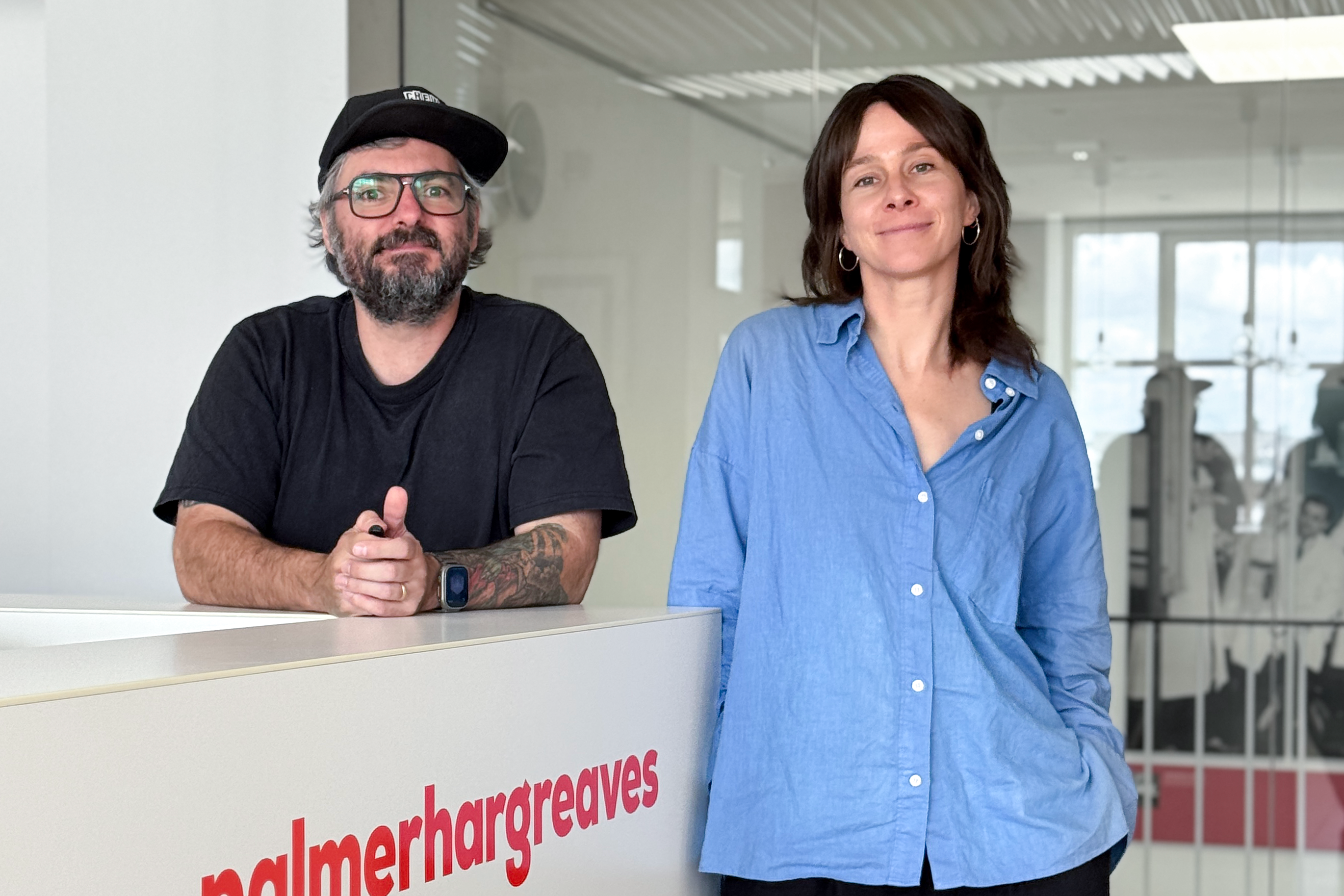 Markus steht links und trägt ein schwarzes T-Shirt sowie eine schwarze Kappe. Mirja steht rechts und trägt eine blaue Bluse. Beide stehen an einem weißen Empfangstresen mit rotem „palmerhargreaves“-Logo in einem modernen Büro mit Glasfront und hellem Interieur.
