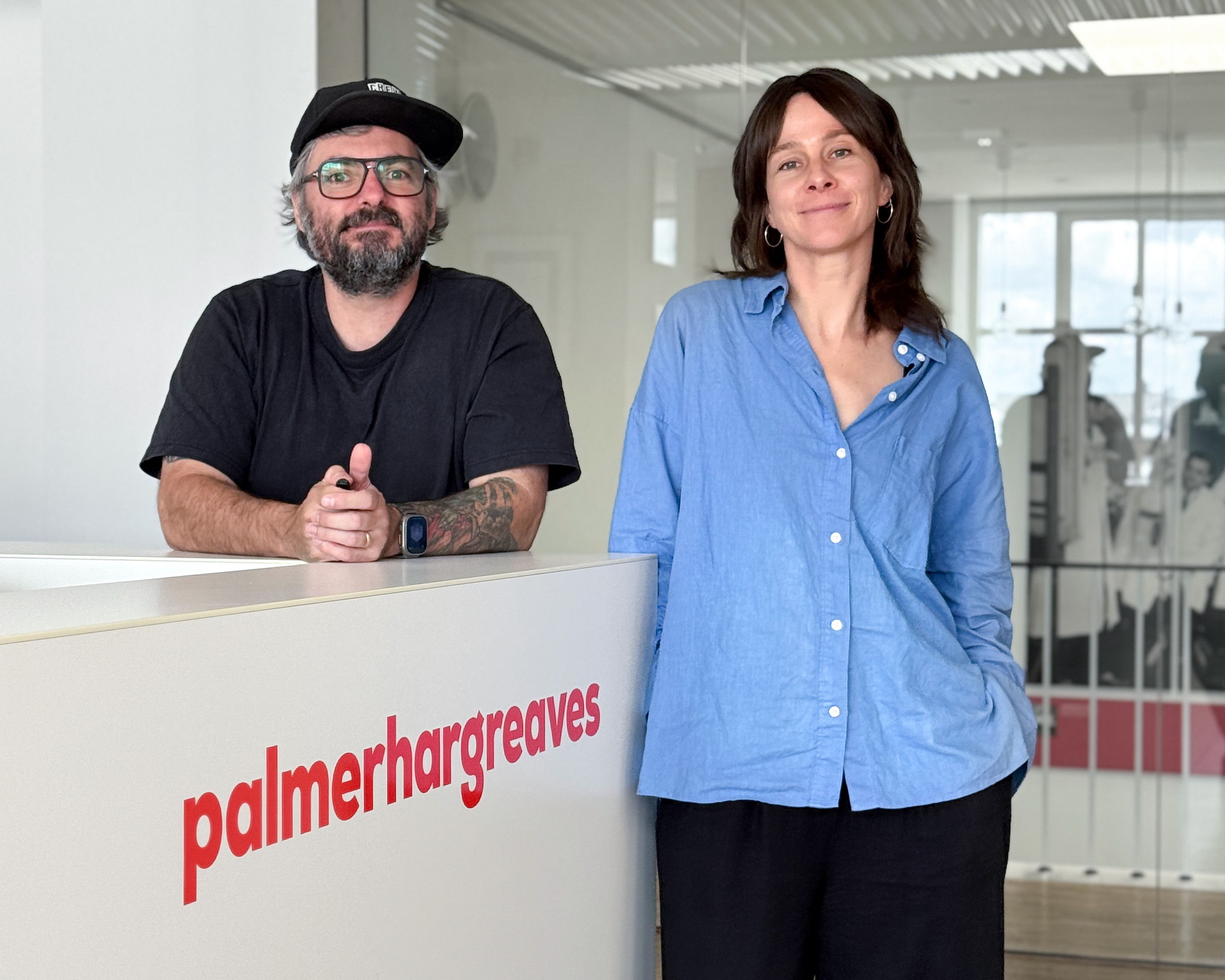 Markus steht links und trägt ein schwarzes T-Shirt sowie eine schwarze Kappe. Mirja steht rechts und trägt eine blaue Bluse. Beide stehen an einem weißen Empfangstresen mit rotem „palmerhargreaves“-Logo in einem modernen Büro mit Glasfront und hellem Interieur.