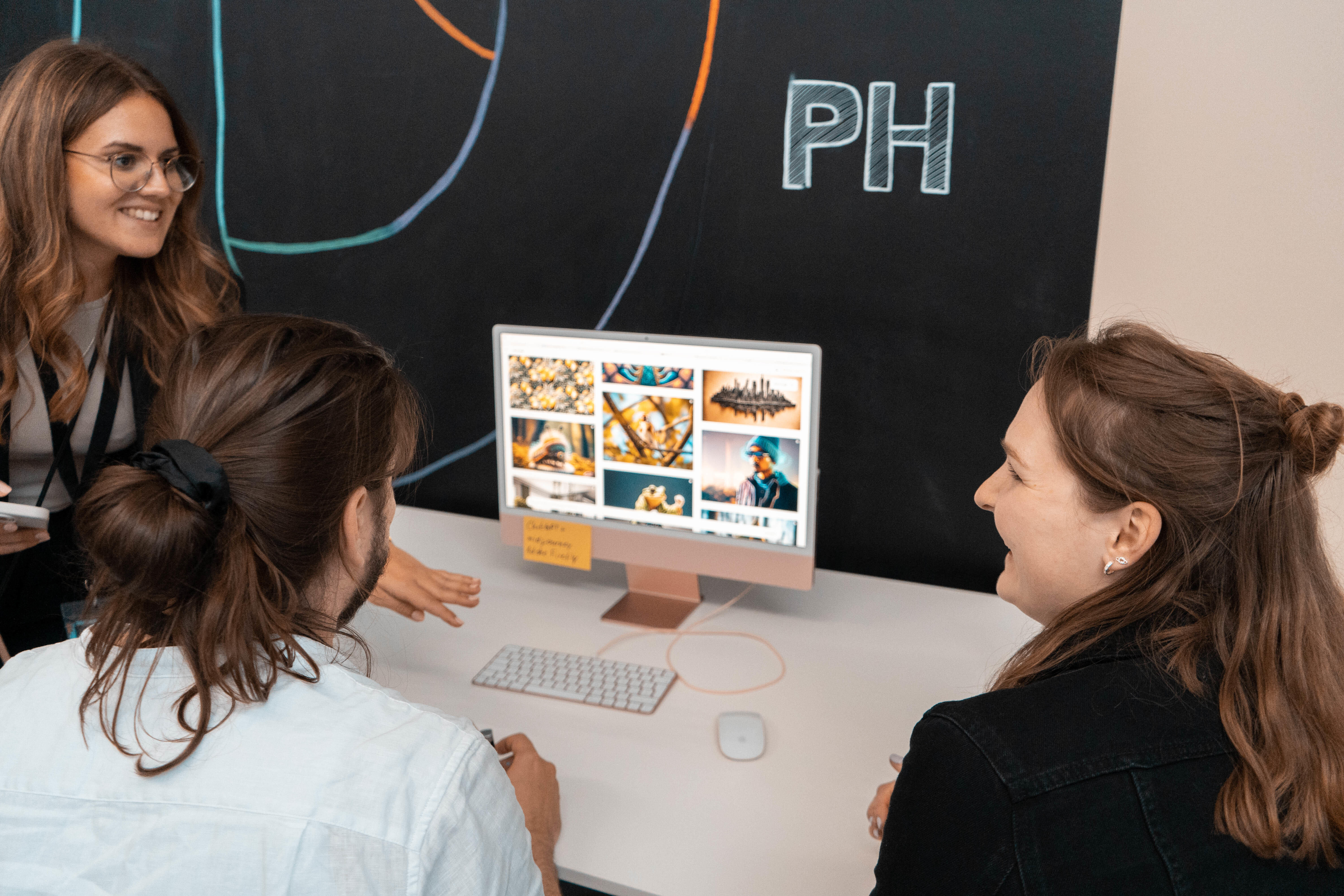 Drei Personen sitzen gemeinsam an einem Schreibtisch und betrachten einen Computerbildschirm mit verschiedenen Bildern, vor einer Wand mit der Aufschrift "PH".