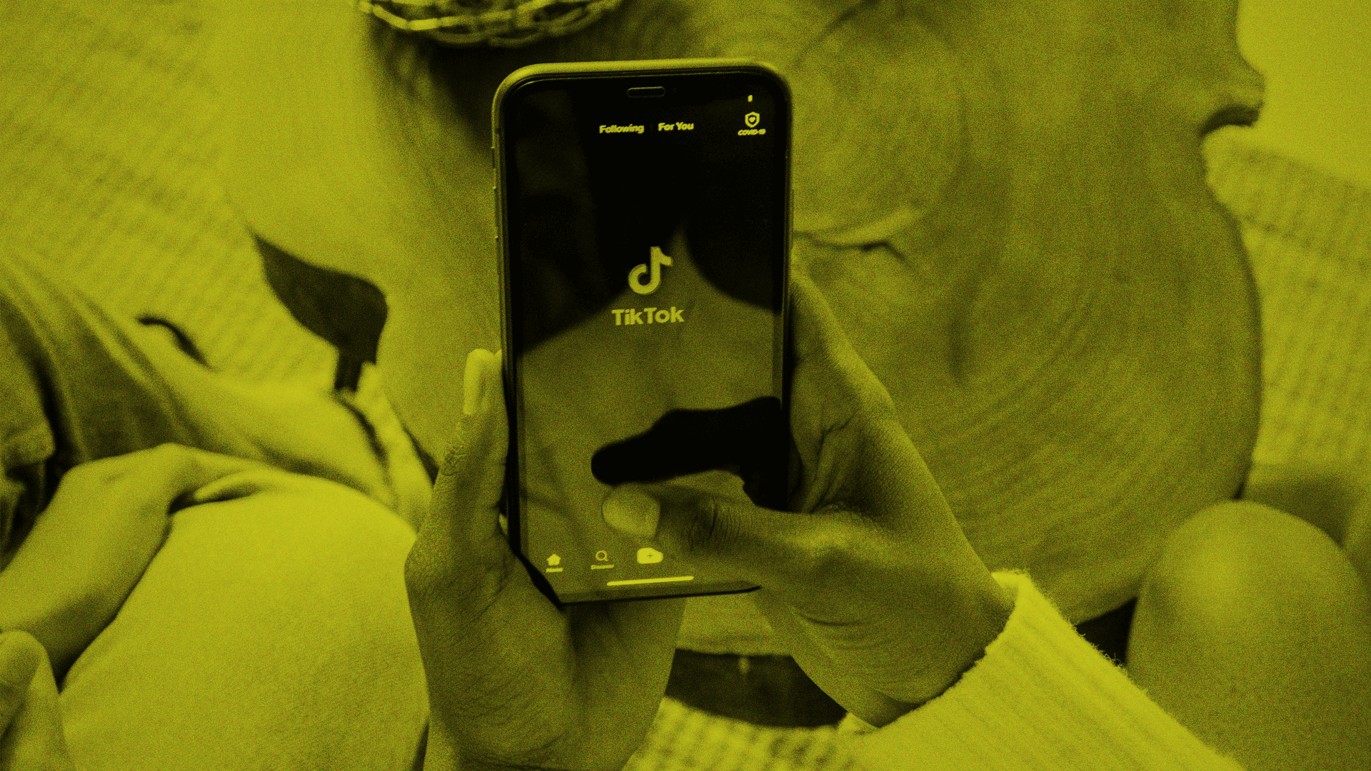 Eine Person hält ein Smartphone mit geöffnetem TikTok-Startbildschirm in der Hand, während im Hintergrund eine weitere Person sitzt. Das Bild ist gelb eingefärbt.