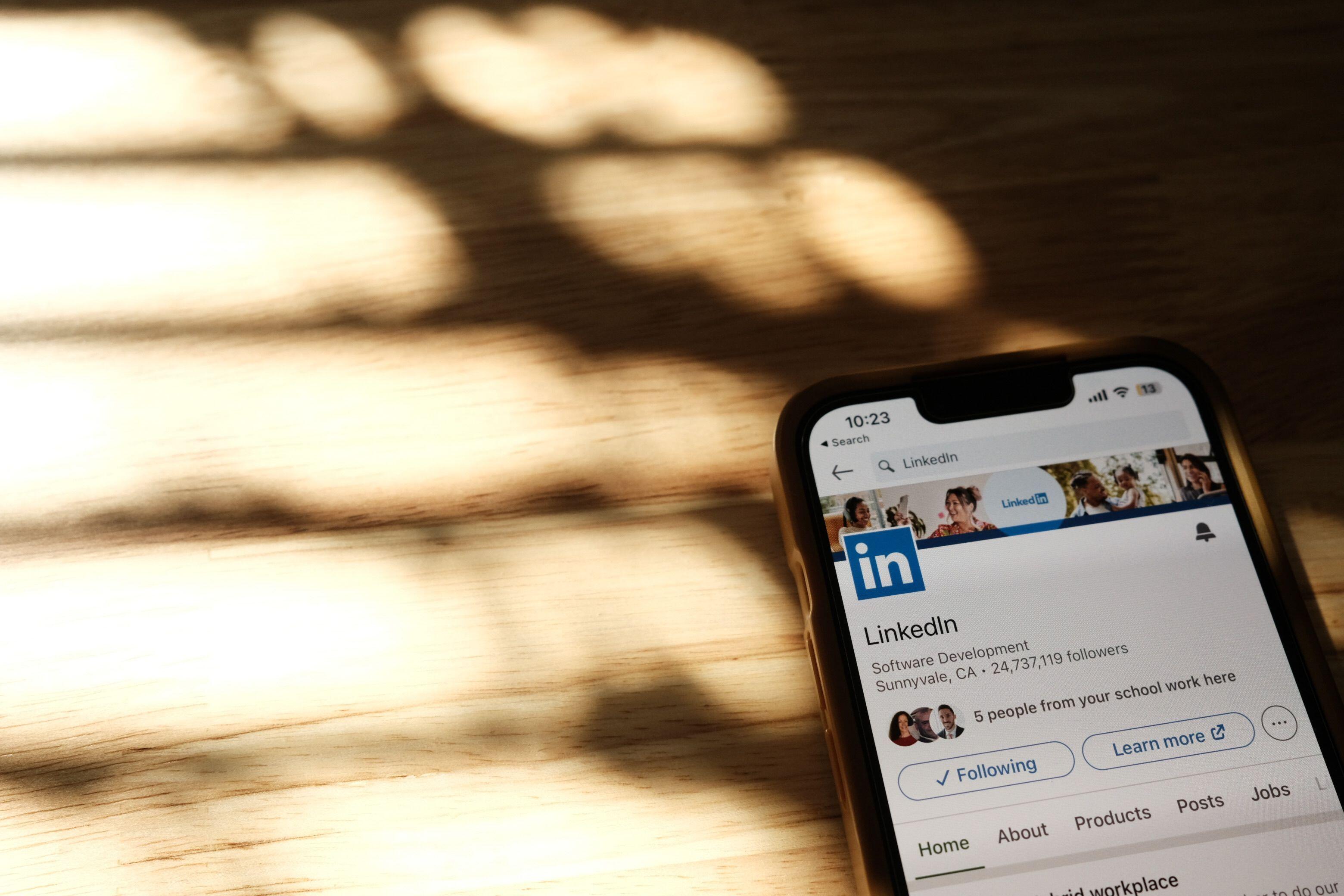 Ein Smartphone liegt schräg auf einem Holztisch, auf dem Sonnenlicht durch Fensterlamellen warme Schattenmuster wirft. Auf dem Display ist die LinkedIn-Profilseite „Linkedin“ geöffnet, mit dem Hinweis „Software Development, Sunnyvale, CA · 24.737.119 followers“ und einem Banner mit Personenfotos. Darunter stehen die Schaltflächen „Following“ und „Learn more“.