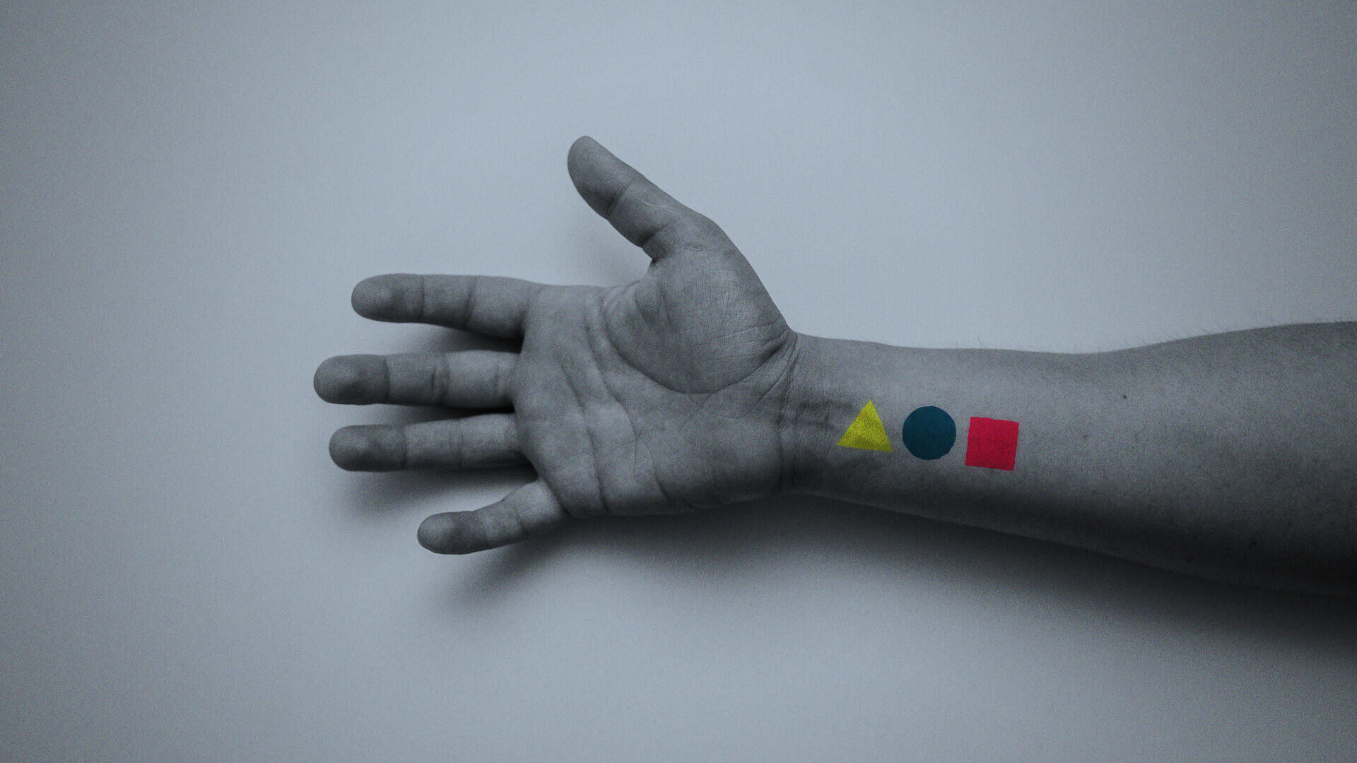 Ein ausgestreckter Arm auf hellem Hintergrund, auf dem Unterarm sind drei farbige geometrische Formen zu sehen: ein gelbes Dreieck, ein blauer Kreis und ein rotes Quadrat.