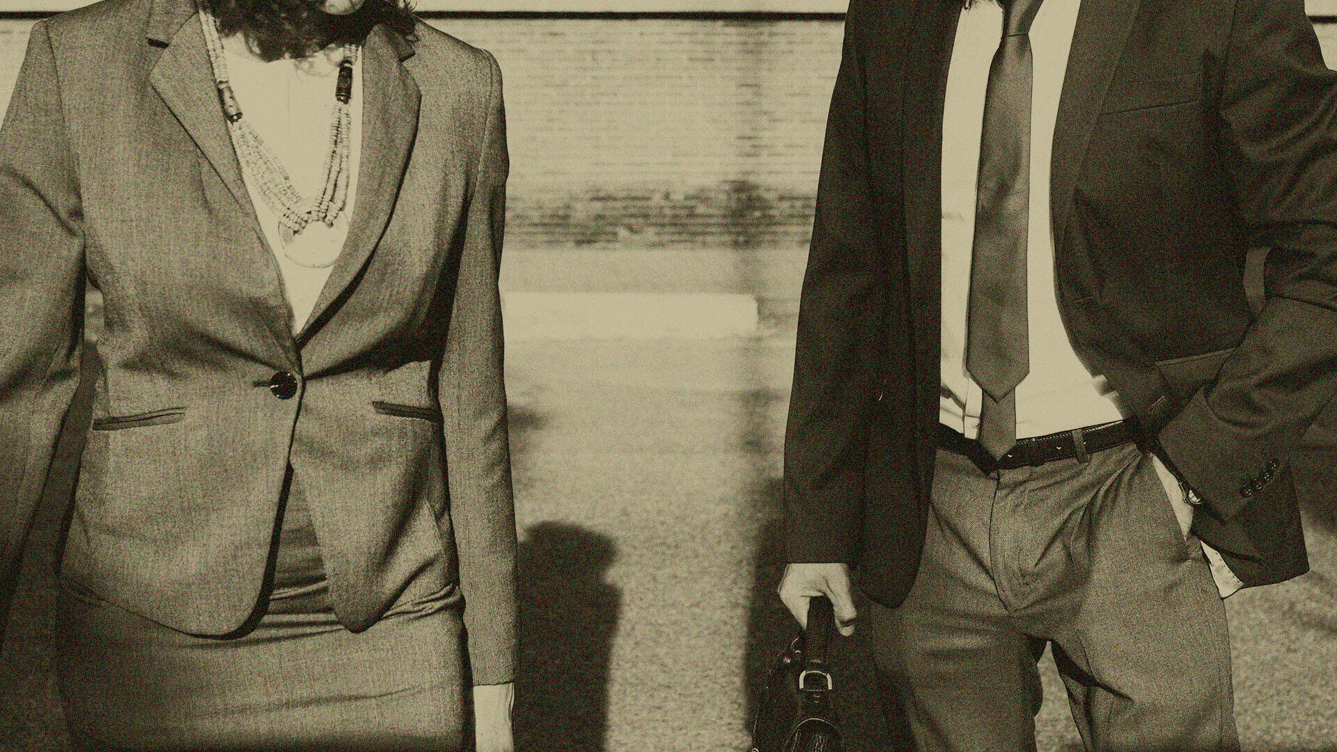 Zwei Personen in Business-Outfits gehen nebeneinander auf einer Straße. Eine trägt einen Rock und Blazer mit Schmuck, die andere einen Anzug mit Krawatte und hält eine Aktentasche. Das Bild wirkt wie eine Vintage-Aufnahme.