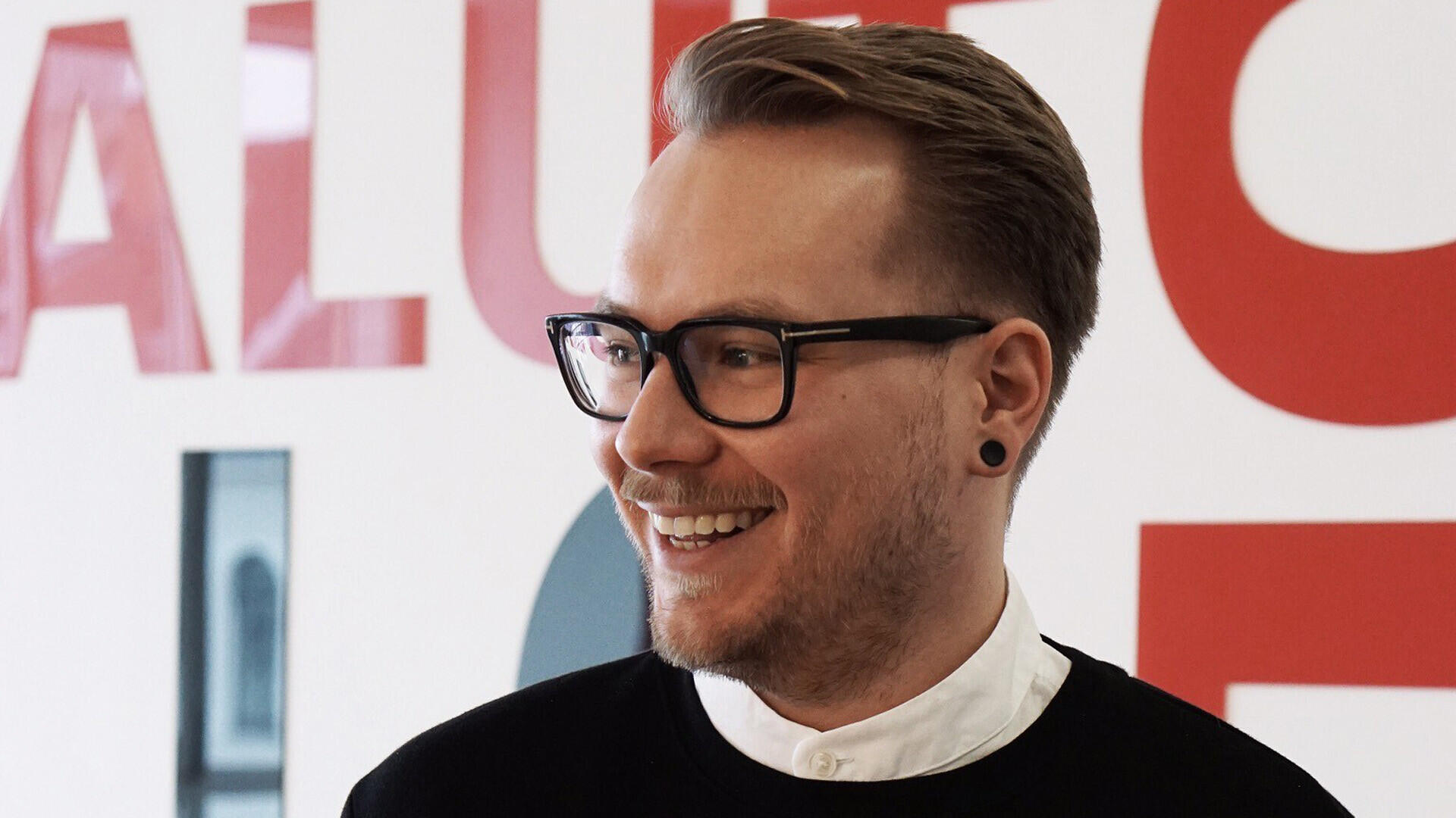 Person mit kurzen, seitlich gestylten Haaren, Brille und Ohrpiercing steht vor einer Wand mit großflächigen roten Buchstaben. Die Person trägt ein weißes Hemd unter einem dunklen Pullover.