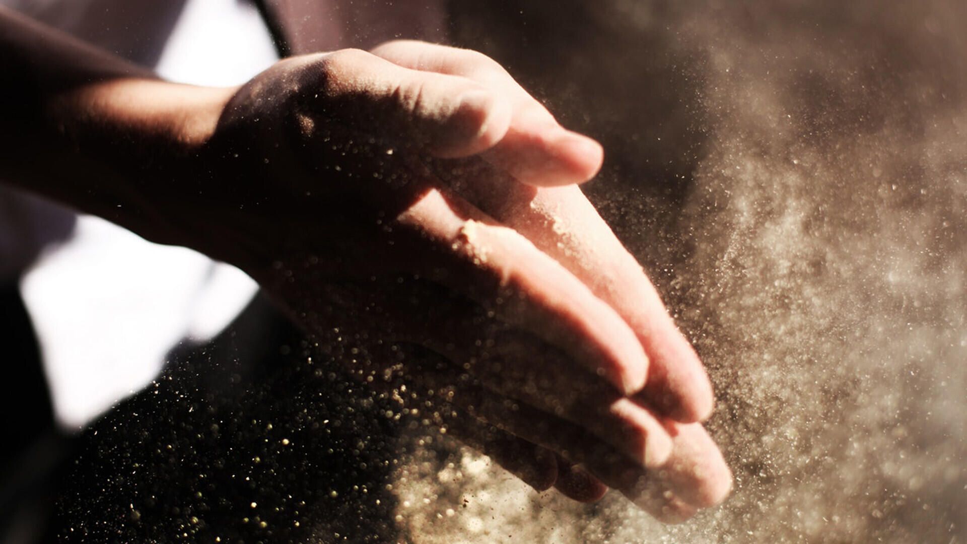 Zwei Hände klatschen zusammen, dabei wirbelt feiner Staub oder Mehl in der Luft auf und wird von Lichtstrahlen beleuchtet.