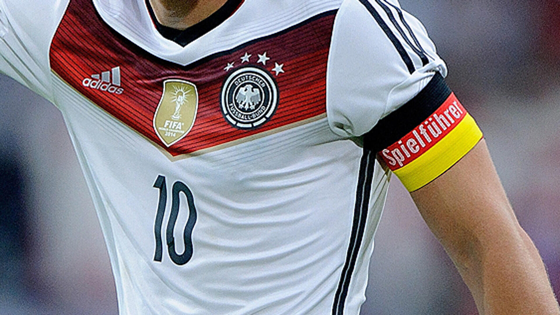 Oberkörper eines Fußballspielers im weißen Trikot der deutschen Nationalmannschaft mit der Nummer 10, Weltmeisterschafts-Emblem und Kapitänsbinde mit der Aufschrift „Spielführer“ am Arm.