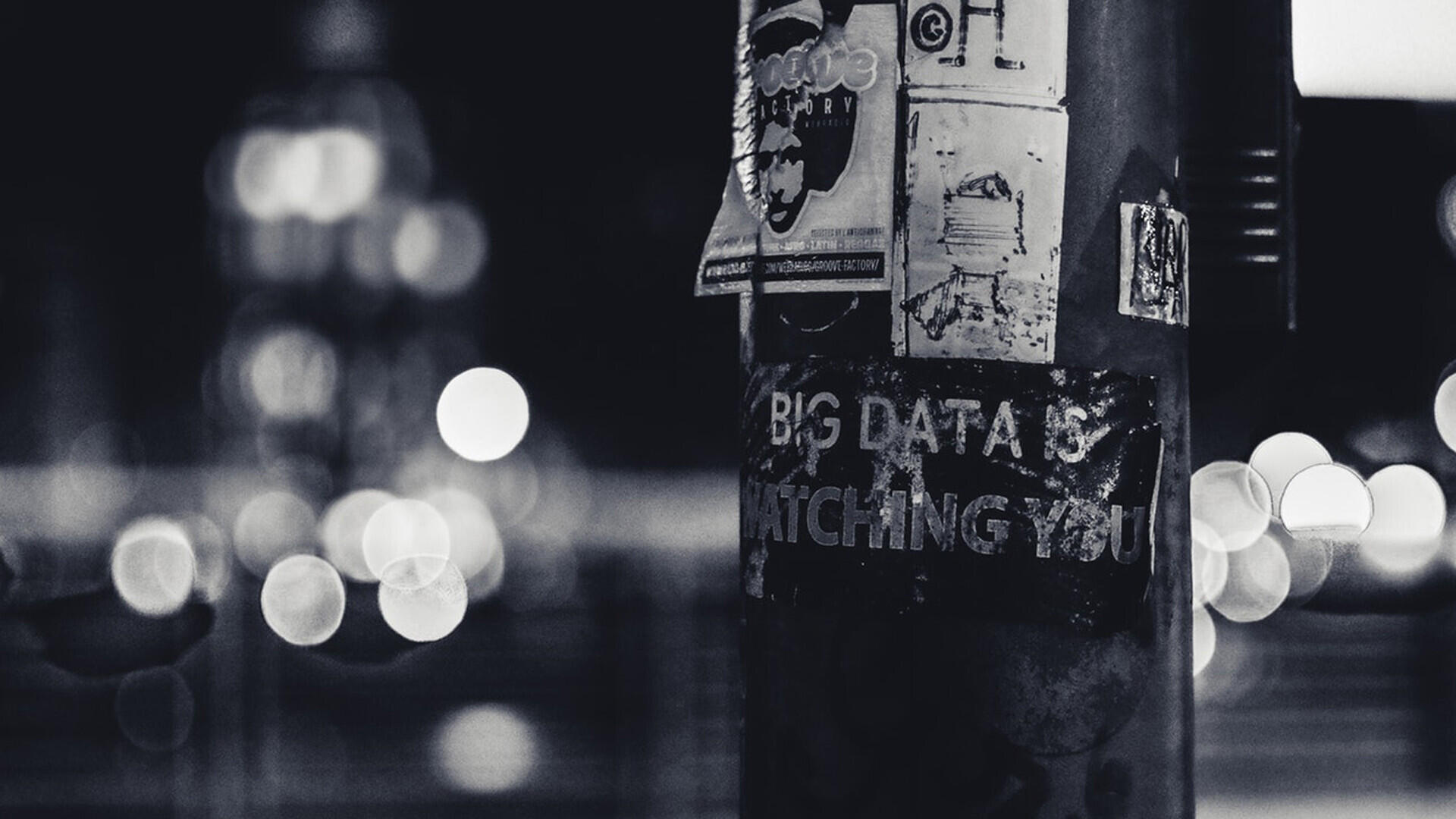 Schwarz-weiß-Foto eines Laternenpfostens mit mehreren Aufklebern, darunter einer mit der Aufschrift „Big Data is watching you“. Im Hintergrund sind unscharfe Lichter einer nächtlichen Stadt zu sehen.