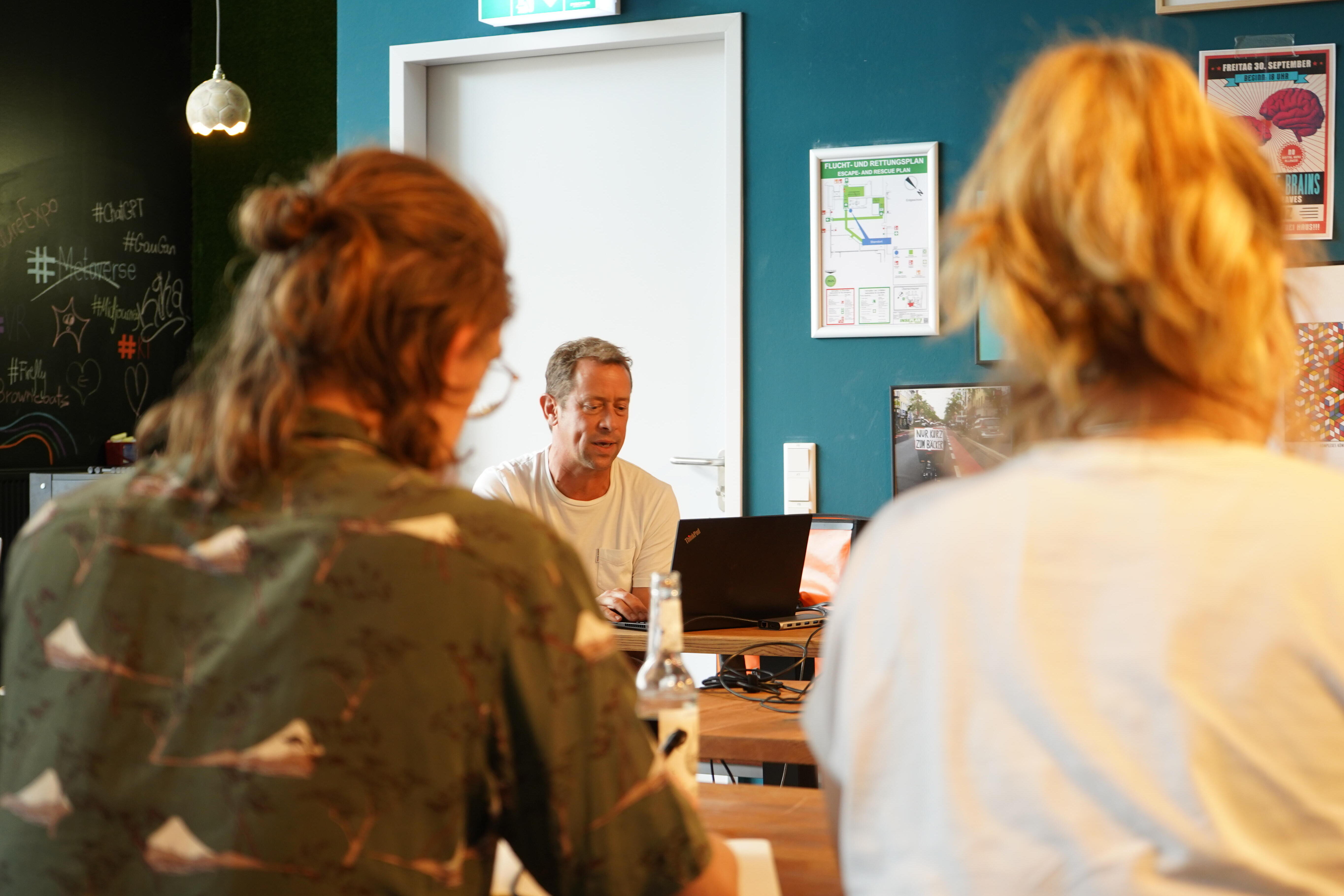 Drei Personen sitzen an einem Holztisch in einem modernen Büro. Im Hintergrund ist eine blaue Wand mit verschiedenen Postern, einem Notfallplan und einer Tür zu sehen. Links befindet sich eine Wand mit Kreidezeichnungen und Hashtags. Auf dem Tisch liegen Laptops und Getränke, die Atmosphäre wirkt entspannt und kreativ.