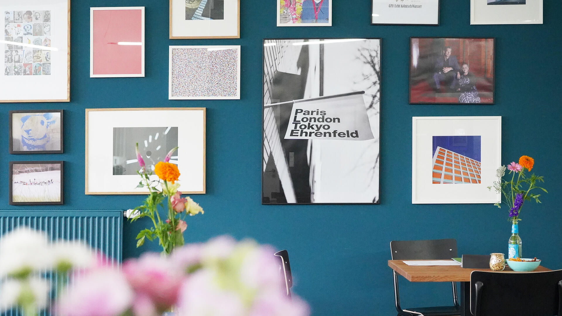 Bunte, modern eingerichtete Sitzecke mit dunkler Wand, an der verschiedene gerahmte Bilder und Fotografien hängen. Auf einem Tisch stehen Blumen in Vasen, daneben Stühle und kleine Dekorationsgegenstände. Im Mittelpunkt ein großes Bild mit der Aufschrift „Paris London Tokyo Ehrenfeld“.