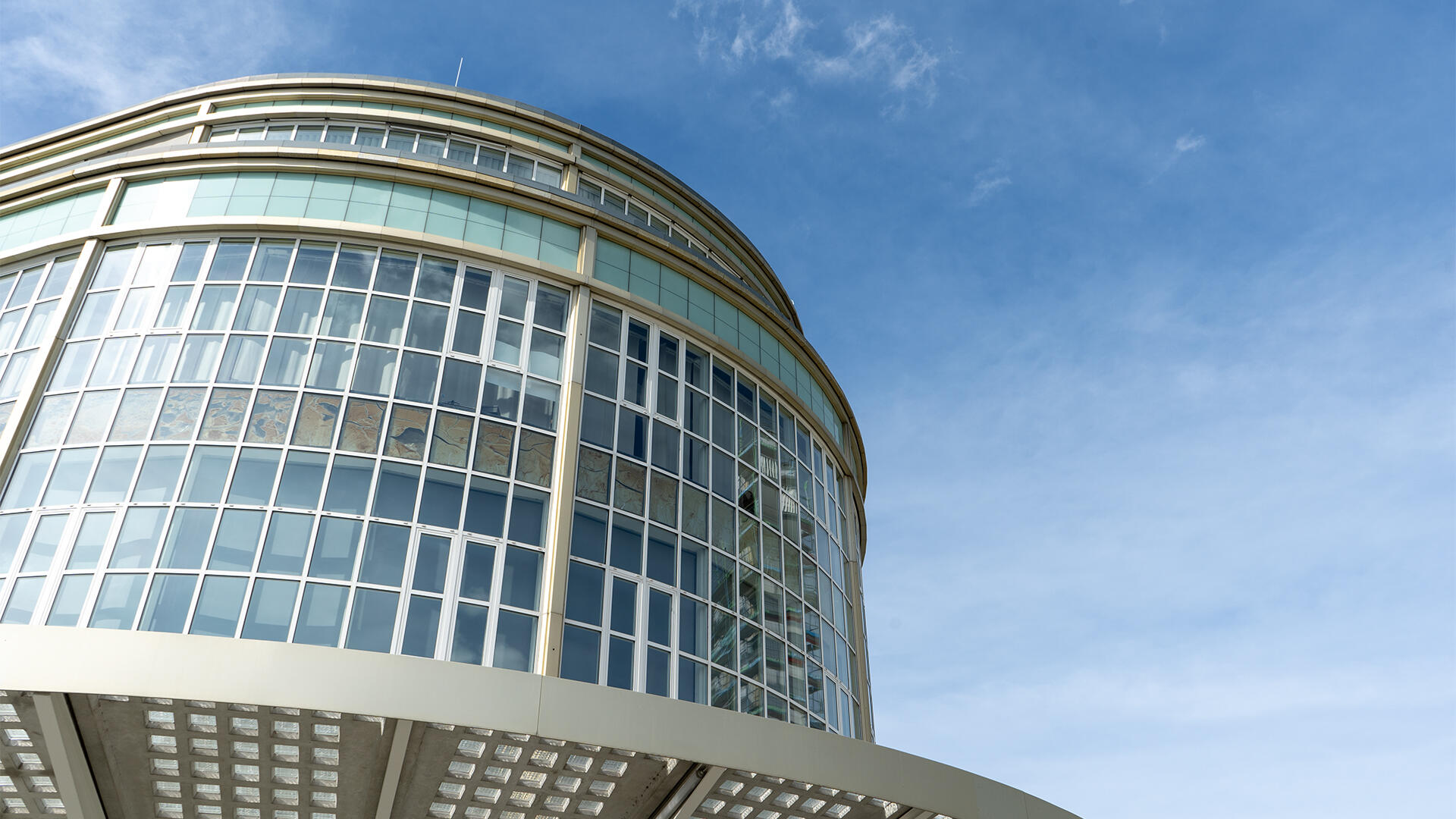 Modernes, rundes Gebäude mit großen Glasfenstern und Rahmen aus Metall, fotografiert aus der Froschperspektive vor blauem Himmel mit wenigen Wolken.