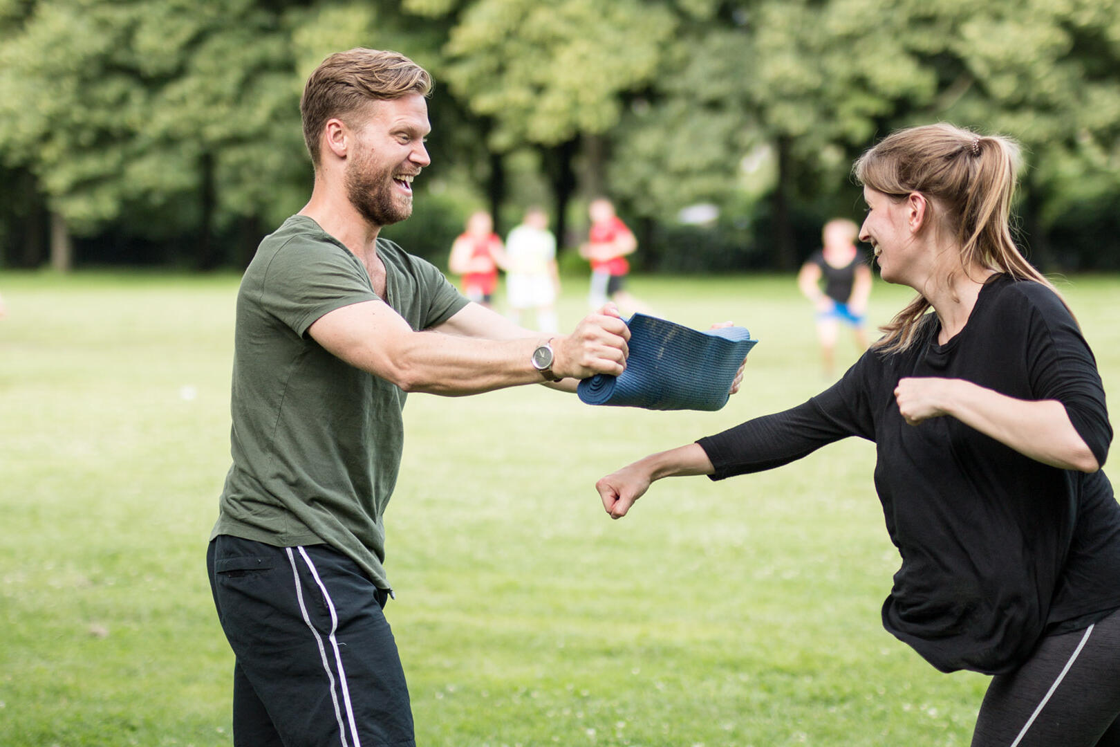 Zwei Personen üben eine Schlagtechnik auf einer Wiese im Park. Eine Person hält ein Schlagpolster, die andere führt einen Faustschlag aus. Im Hintergrund sind weitere Menschen und Bäume zu sehen.